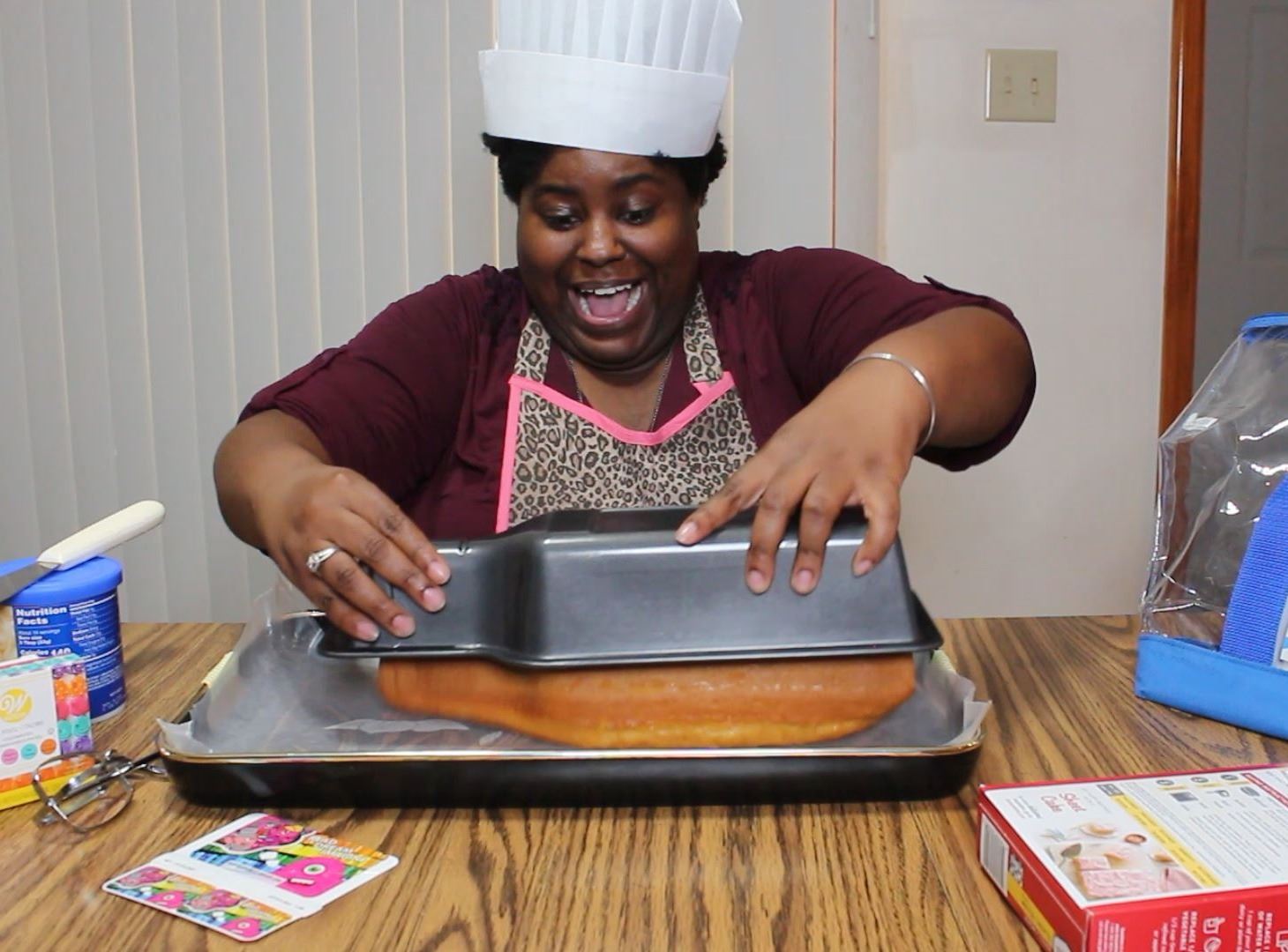 Library staff wearing chefs hat taking bottle shaped cake out of pan at table